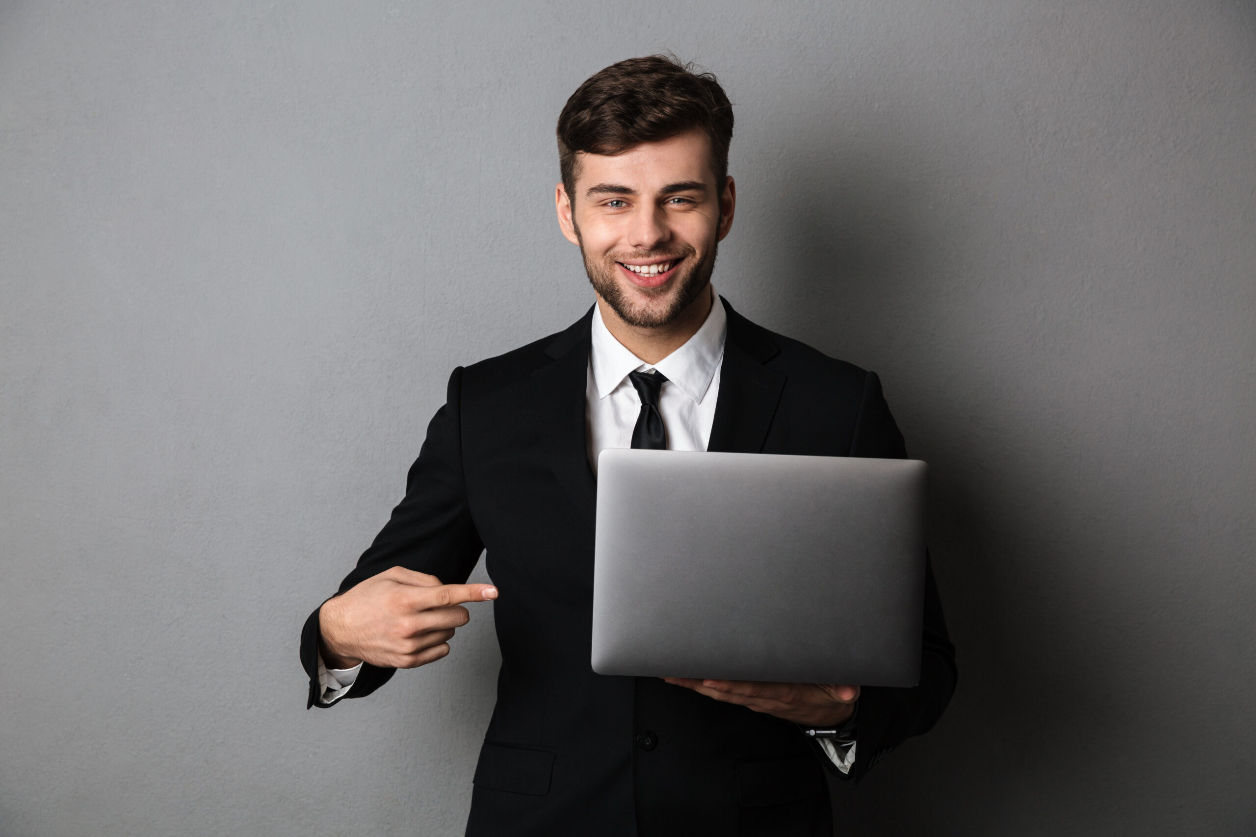 Close-up portrait of cheerful businessman pointing with finger on his laptop computer, isolated on gray background